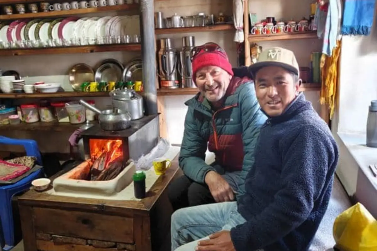 A smiling trekker and local guide sitting by a warm indoor wood-burning stove during the Annapurna Circuit Trek Tour.