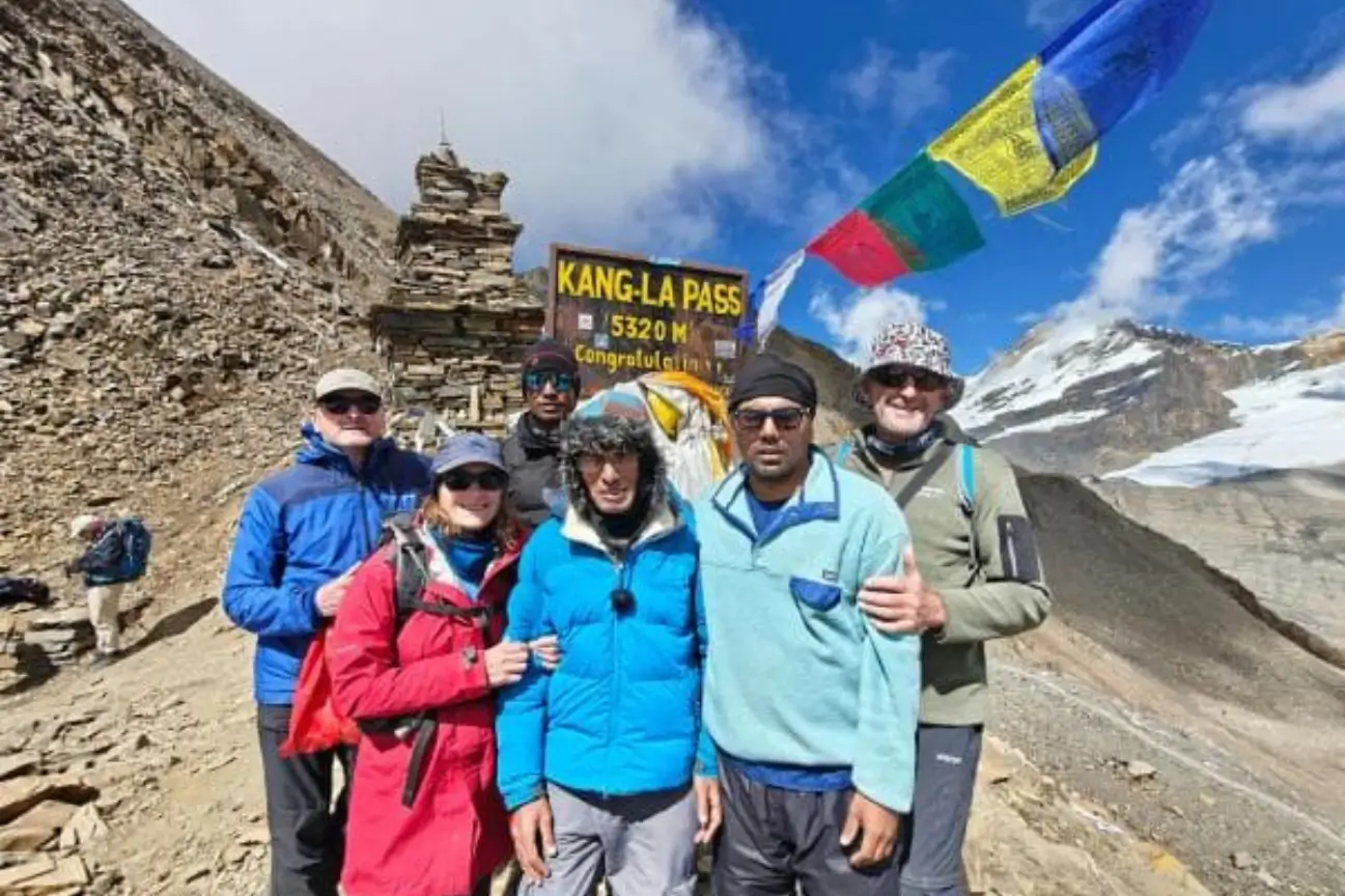A group of trekkers and local guides posing by a milestone sign and prayer flags at Kang-La Pass during the Annapurna Circuit Trek Tour.