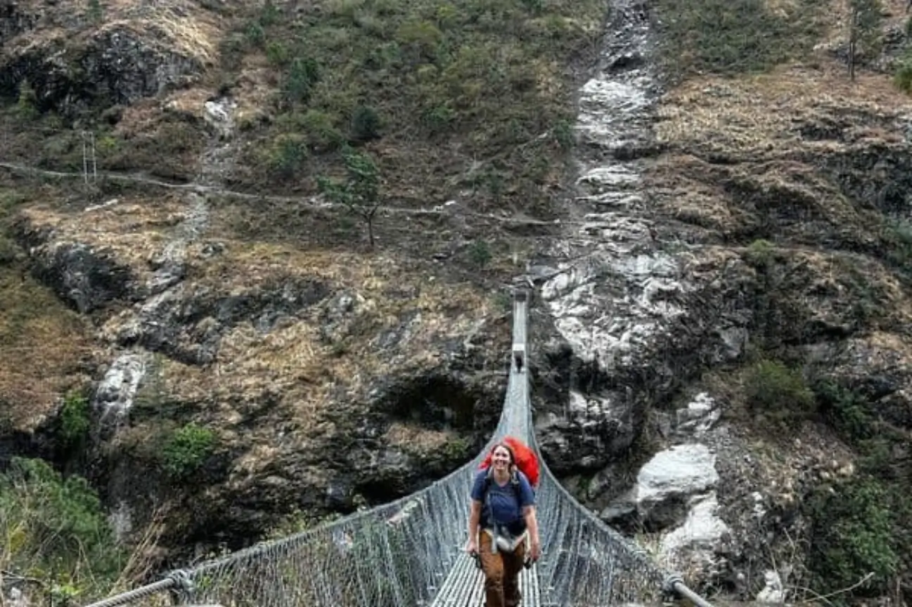 A smiling trekker with a red backpack walking across a long suspension bridge on the Annapurna Circuit Trek Tour.